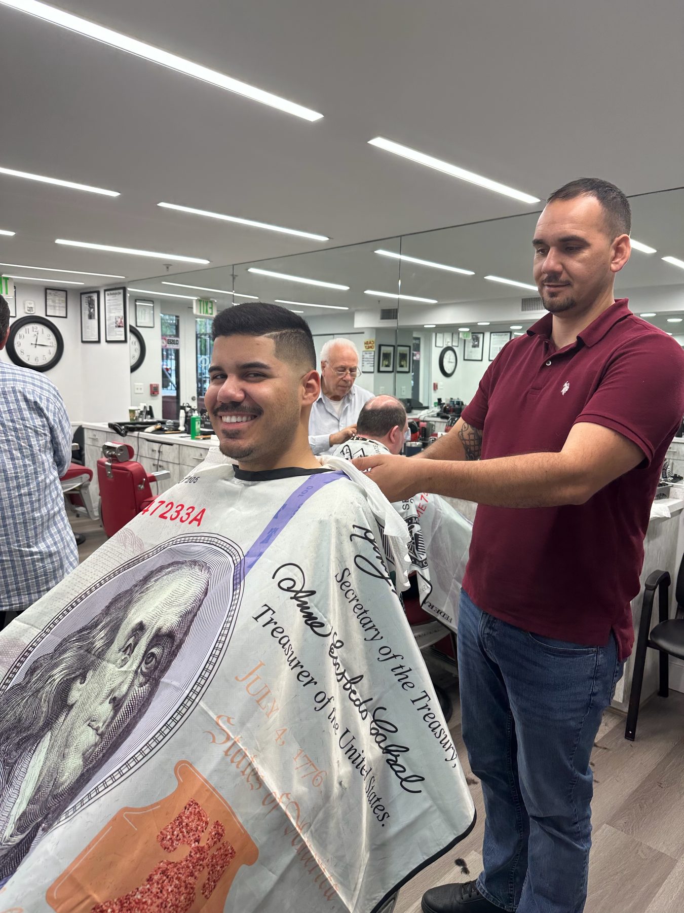 Um barbeiro a acabar um corte enquanto o Tony trabalha numa cadeira, refletido no espelho